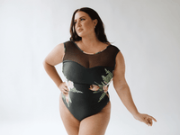 Front view of a black floral one-piece swimsuit with sheer mesh neckline on a model against a light background