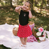 Young girl standing on a picnic blanket wearing a cute kid's Disney outfit—kids Minnie tank top and kids tennis skirt in red polka dots.