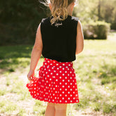 Back view of young girl wearing a kids Minnie tank top and kids tennis skirt for Disneyland on a blurred greenery background.