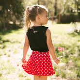 Back view of young girl standing in a park wearing a red polka dot Disney tennis skirt for kids with a black Minnie Mouse tank top.