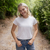 A smiling blonde woman wearing a plain white short-sleeve crewneck t-shirt and blue jeans, standing on a dirt path surrounded by green trees.