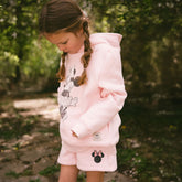 Child wearing a pink Minnie hoodie and matching kids Minnie shorts in pink on a blurred greenery background.