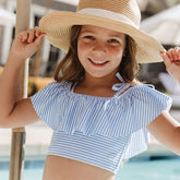 Young girl wearing a blue and white striped swimsuit top and wide-brimmed hat by a pool.