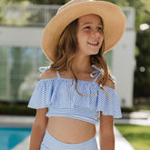 Young girl wearing a blue and white striped little girls swimsuit with a straw hat by a pool.