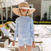 Child wearing a blue striped long sleeve kids swimsuit and wide-brimmed hat by a poolside.