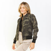 A woman wearing a green camouflage bomber jacket with gold zipper details and an embroidered logo on the chest.