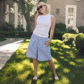 A woman wearing a white sleeveless top and light blue striped knee-length shorts stands on a sunny grassy lawn in front of a house.