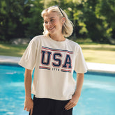 Woman wearing a 'USA 1776' t-shirt by a pool with greenery in the background