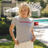 Woman wearing a striped t-shirt with 'america' printed on it, standing outdoors near a pool.