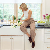 Woman sitting on a kitchen counter arranging flowers, wearing women's balloon pants in brown gingham print with a cropped white tank top.