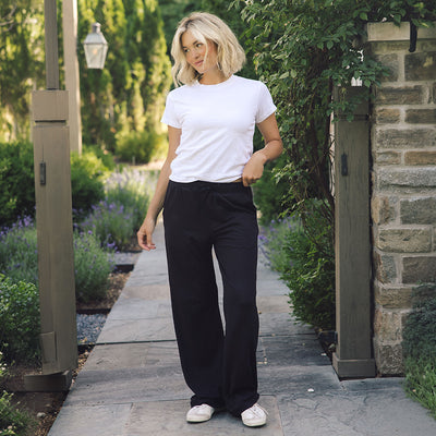 A woman stands on a stone walkway outdoors wearing a white short-sleeve crewneck t-shirt, loose black wide-leg pants, and white sneakers.