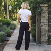 A woman viewed from behind wearing a white short-sleeve crop top and black wide-leg pants, standing on a stone pathway in a garden setting next to a stone pillar.