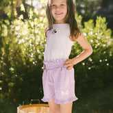 Young girl wearing a white tank top and purple checkered shorts with Minnie Mouse details on a blurred outdoor setting.