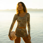 Woman in a long sleeve rash guard swimsuit top with matching boy short swim bottoms holding a large seashell on a beach