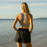 Back view of woman wearing a black and white striped criss cross back swim top and black linen shorts standing by a body of water.