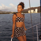 Woman in a polka dot swimsuit standing on a boat with water and mountains in the background