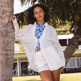 Woman in a white shirt and shorts cover-up set over blue floral swimsuit leaning against a palm tree.