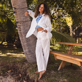 Woman in a white cotton pants and flowy shirt over a swimsuit standing next to a palm tree in a tropical setting