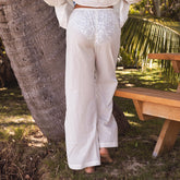 Back view of woman wearing white cover up pants over a swimsuit standing next to a wooden bench