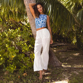 Woman in a blue floral tankini top and white linen pants leaning against a tree in a tropical setting