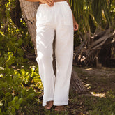 Woman wearing white linen pants as a beach cover-up standing in a natural setting with greenery.