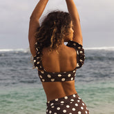 Woman in a polka dot swimsuit standing by the ocean with arms raised.
