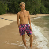 Man wearing pink floral swim shorts standing on a beach with waves and greenery in the background