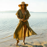 Woman in an olive green wrap cover-up over trendy bikini with a straw hat on standing on a beach.