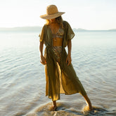 Woman in a long green cover-up dress over a stylish bikini and straw hat standing in shallow water.