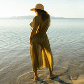 Back view of woman in a long green cover-up dress and hat standing in shallow water at sunset.