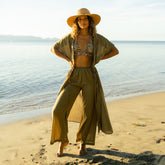 Woman in two piece swimsuit with olive green beach cover up pants and beach wrap standing on a beach.