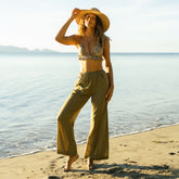 Woman in a a bikini top and olive green beach pants standing on a beach with ocean in the background
