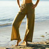 Woman wearing olive green, wide-leg pants standing on a beach with water in the background