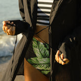 Close up of woman wearing a black cover-up top over a green leaf print swimsuit on a beach.