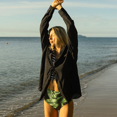 Woman stretching her arms above her head on a beach, wearing a black and white bikini top, palm green high waisted bottoms, and a black wrap cover up top.