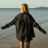 Back view of woman wearing a black button down shirt standing by the water with arms outstretched