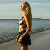 Woman in a striped swimsuit top and black cotton shorts standing on a beach with ocean in the background
