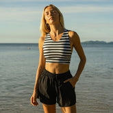 Woman in a striped swimsuit top and black cover up shorts standing on a beach with water and sky in the background.