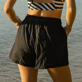 Back view of woman wearing a black and white striped bikini top and black cover-up shorts by a body of water