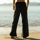 Back view of woman wearing black wide-leg cover up pants standing on a beach with water in the background