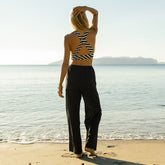 Back view of woman standing on a beach wearing a striped swim top and black linen pants as a cover-up, with a scenic ocean view.
