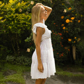 Side view of woman in a short sleeved white dress standing in a garden with flowers and greenery.