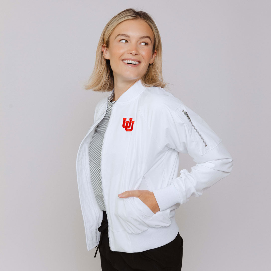 A woman smiling, wearing a white bomber jacket with the University of Utah logo on the chest, paired with black pants.