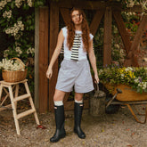 Model standing in front of a shed holding a watering can. She's wearing blue seersucker women's shorts and hunter boots.