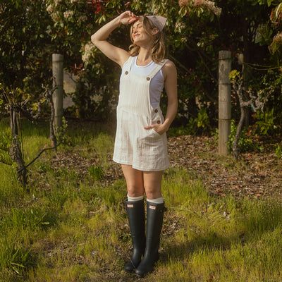 Model wearing white cotton short overalls with brown buttons over a white tank top with black rain boots and a gingham headscarf.