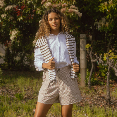 Model in a vineyard wearing preppy women's shorts in brown stripes with a white button down shirt and a sweater over her shoulders.