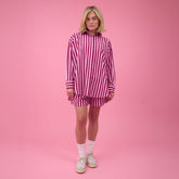 Woman wearing a pink striped matching set—oversized button down shirt and boxer shorts—in a studio with a pink background.