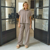 Woman in a brown and cream striped matching set, a short sleeved button down and striped trousers standing in a modern interior.