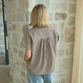 Back view of woman wearing a striped, short sleeved button down shirt and jeans standing against a stone wall.