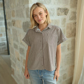 Woman wearing a short sleeve button down shirt in brown and cream stripe with blue jeans standing against a stone wall.